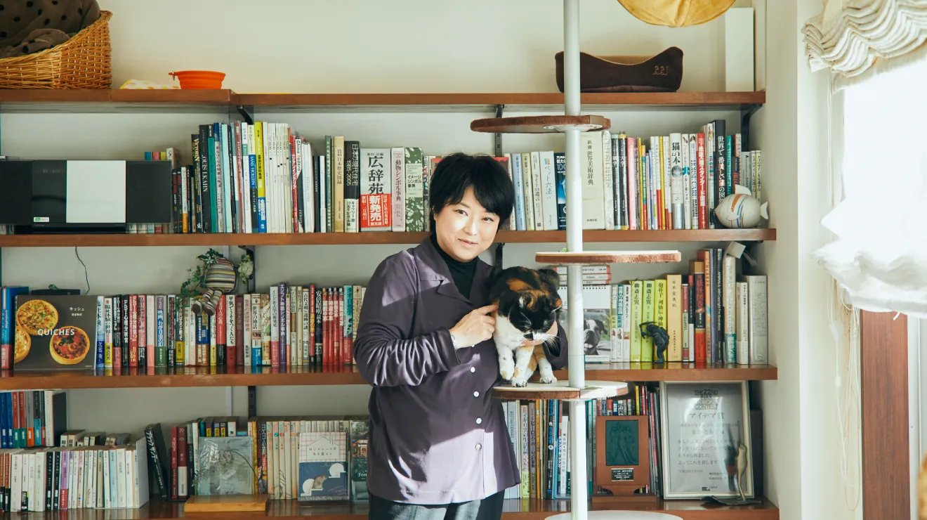 本棚の前で猫と一緒に立つ一級建築士・金巻とも子さん