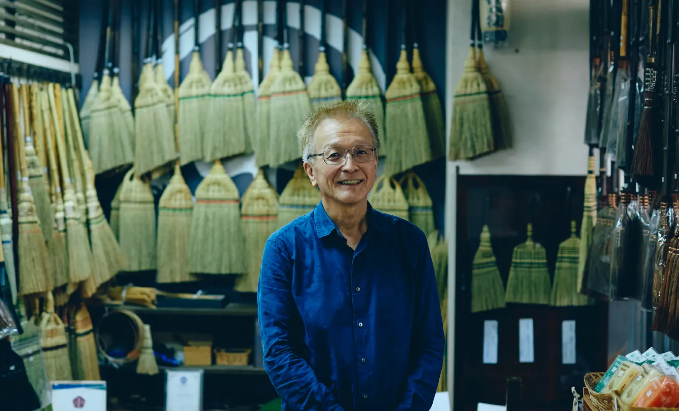 店内で江戸箒を背に笑顔を見せる中村悟さん